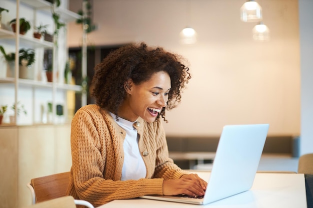 Agradable joven mujer étnica sonríe mientras escribe texto en el teclado de la computadora portátil teletrabajando en la oficina