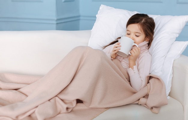 Agradable niña enferma que sufre de gripe y bebe té mientras está acostada en su mal