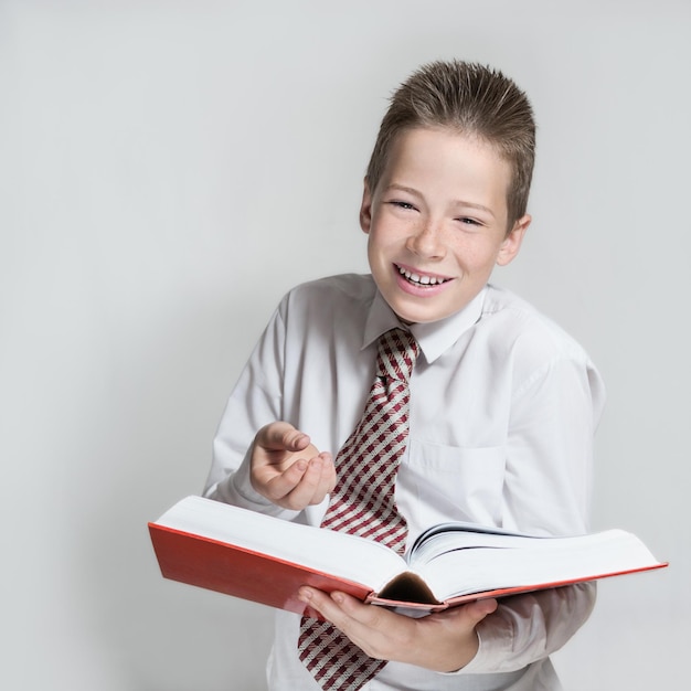 El agradable niño adolescente sonriente con una camisa blanca y una corbata lee un gran libro rojo divertido de fondo gris