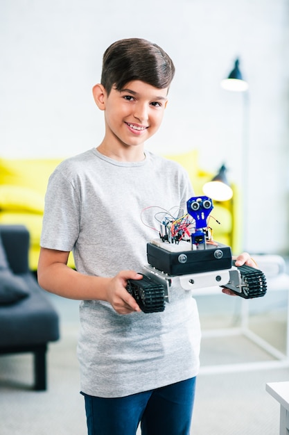 Agradable niño inteligente sonriente sosteniendo un robot moderno hecho por él para la competencia de ingeniería