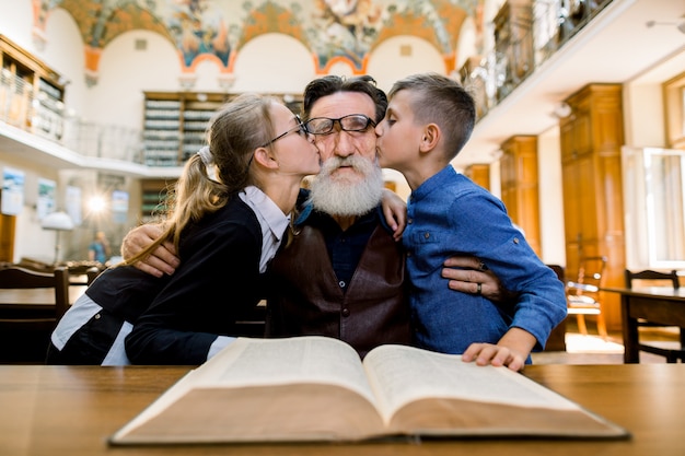 Agradável avô barbudo passando um tempo na biblioteca com a neta e o neto, beijando-o nas bochechas