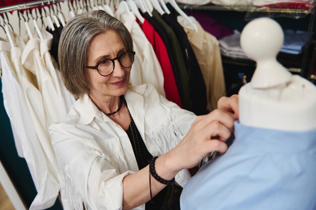 Agradável designer de moda feminina madura experimentando camisa azul no modelo de uma costureira em seu ateliê de alfaiataria preparando nova coleção de roupas exclusivas para exposição