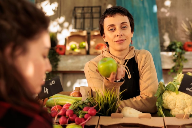 Agradável jovem agricultora em pé atrás do mercado, com produtos orgânicos saudáveis, segurando maçã verde e sorrindo para a câmera. Mulher vendendo frutas e legumes frescos cultivados localmente