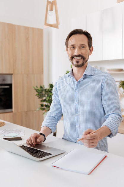 Agradável jovem barbudo em pé atrás do balcão da cozinha, segurando uma mão no laptop enquanto pega um lápis na outra enquanto parece feliz