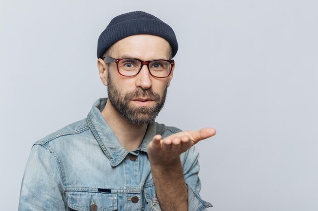 Agradável jovem homem barbudo com bigode usa óculos da moda chapéu preto e camisa jeans sopra beijo de ar na câmera expressa seu amor à namorada isolado sobre fundo cinza