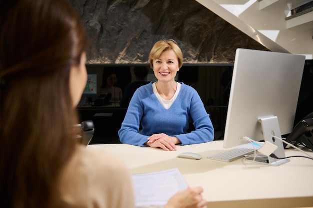 Agradável mulher madura europeia sorrindo sorriso cheio de dentes sentado na mesa em frente a um monitor de computador e conversando com seu cliente no prédio de escritórios moderno