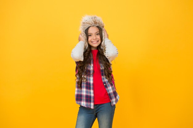 Agregue un poco de personalidad al guardarropa para clima frío. Niño feliz con sombrero de piel sintética. Sonrisa de niña con sombrero de fondo amarillo. Sombrero de trampero esponjoso y cálido para niños. Sombrero de moda para aventuras al aire libre.