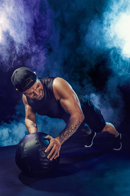 Agressivo desportista musculoso barbudo está malhando, empurre para cima com uma medicine ball isolada na parede escura com fumaça.