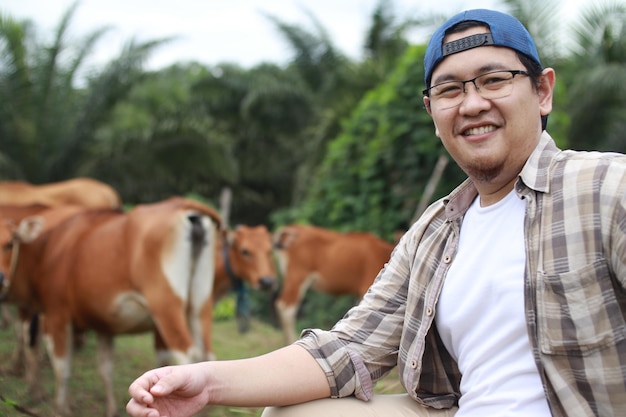 Agricultor asiático feliz e bem sucedido olhando para a câmera e sorrindo com o pastoreio de gado domesticado