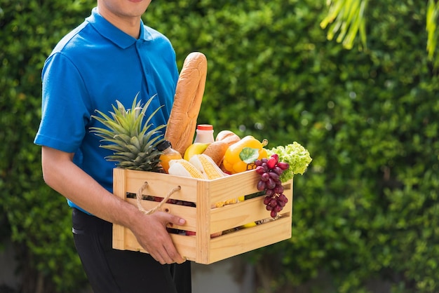 Agricultor asiático usa uniforme de entrega, ele segura frutas e legumes frescos completos em caixa de madeira em mãos, pronta para dar ao cliente colheita de alimentos orgânicos no jardim, fundo de folhas verdes