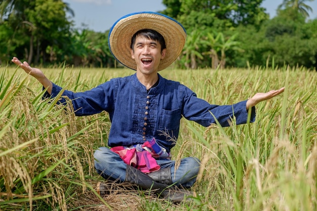 Agricultor asiático usando vestido azul tradicional sentado levantando as mãos com uma cara de uau no meio do campo