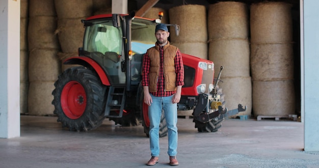 Agricultor bonito de pé em uma camisa e colete laranja sorrindo para a câmera em um fundo de trator e palheiro Alegre trabalhador masculino na fazenda agrícola Máquina para agricultura