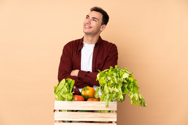 Agricultor com legumes recém colhidos em uma caixa isolada na parede bege, olhando para cima enquanto sorrindo