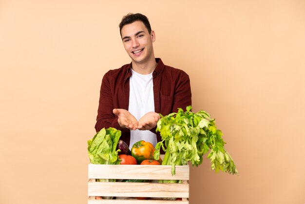 Agricultor com legumes recém colhidos em uma caixa isolada na parede bege segurando copyspace imaginário na palma da mão para inserir um anúncio