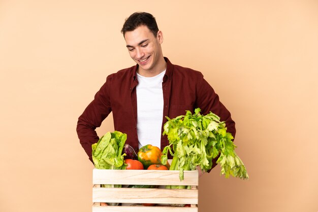 Agricultor com legumes recém colhidos em uma caixa na parede bege posando com os braços no quadril e sorrindo