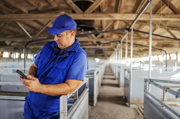 Agricultor com roupa de trabalho em pé e encostado. Ele está usando um smartphone para ligar para o distribuidor de animais