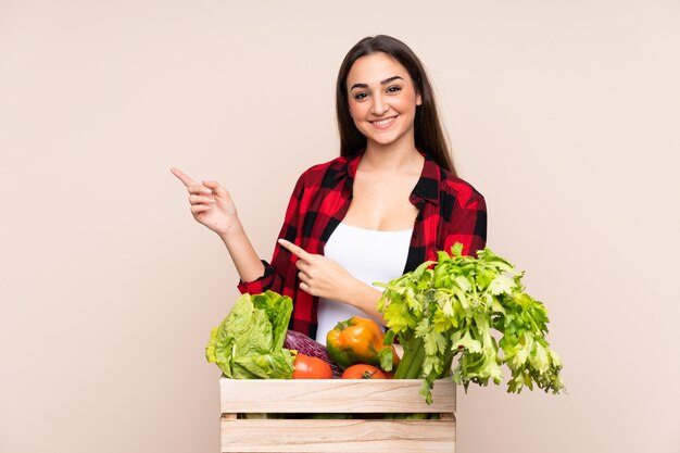 Agricultor com vegetais recém colhidos em uma caixa isolada na parede bege apontando o dedo para o lado