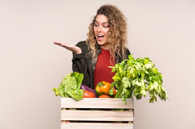 Agricultor com vegetais recém-colhidos em uma caixa isolada na parede bege segurando copyspace imaginário na palma da mão