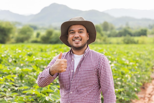 Agricultor feliz com chapéu em pé aparecendo o polegar e sorrindo no campo de plantação ao pôr do sol.