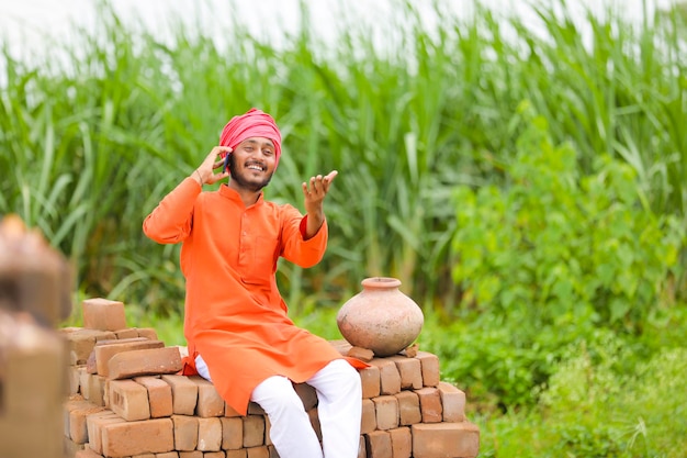 Agricultor indiano sentado em tijolos e falando no smartphone no campo de milho verde.