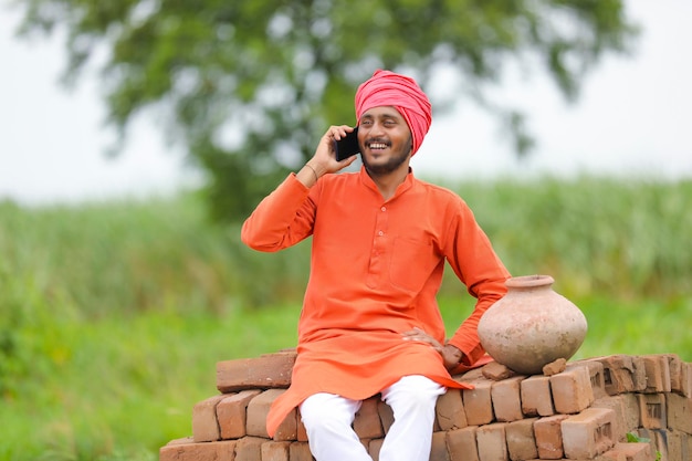 Agricultor indiano sentado em tijolos e falando no smartphone no campo de milho verde.