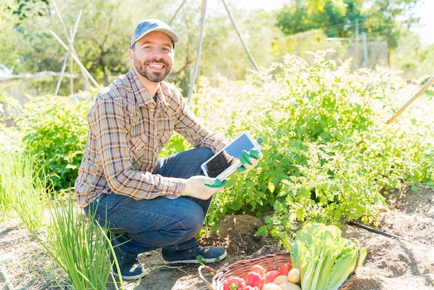 Agricultor latino sonriente usando tableta digital mientras cosecha vegetales en el jardín