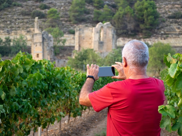 Agricultor mais velho em um vinhedo com um mosteiro em ruínas ao fundo tirando uma foto com seu celular