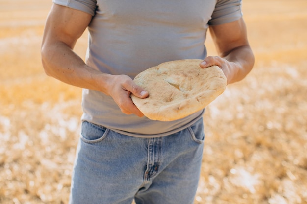 Agricultor masculino em jeans segurando pão fresco perfumado e de pé no campo ensolarado ao redor dos fardos