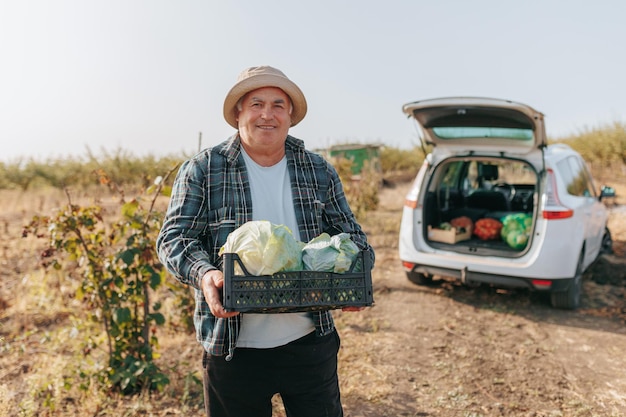 Un agricultor mayor feliz y sonriente mirando a la cámara orgullosamente posa con una caja de repollo fresco y