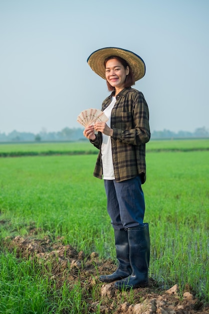 Agricultora asiática vestida con blusas marrones y vaqueros de pie y posa sosteniendo billetes de dinero con una expresión sonriente