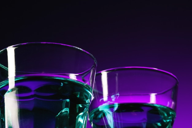 El agua en dos vasos sobre fondo lila en el estudio. Iluminación de colores vivos y brillantes. Bombilla de luz ultravioleta de moda en 2018. Decoración de arte con tono de color místico