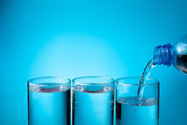 Foto Água em copos, enchendo três copos de uma garrafa de plástico sobre um fundo azul.