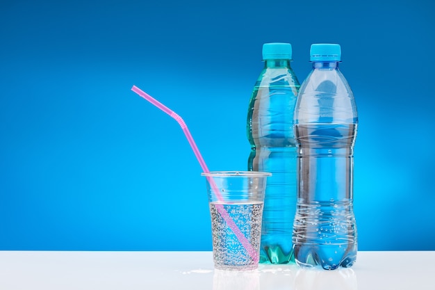 Agua con gas en vaso de plástico con pajita rosa. Botella con agua dulce sobre mesa blanca y fondo azul.