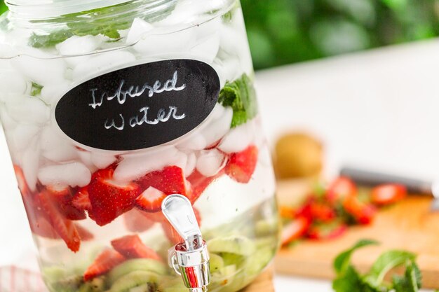 Agua infundida con fresas orgánicas, kiwi y menta fresca en dispensador de bebidas de vidrio.