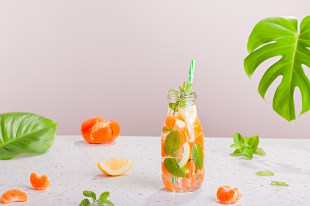 Agua infundida de naranja y menta en botella de vidrio en la mesa Bodegón con cóctel de verano sombrero de paja hoja de monstera Refresco de verano