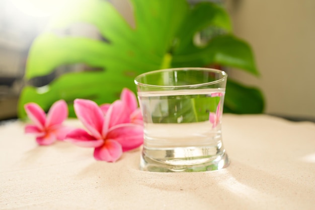 Agua mineral de vidrio en la arena junto a flores de frangipani rosa