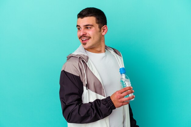 El agua potable del hombre caucásico deportivo joven aislado en azul mira a un lado sonriente, alegre y agradable.