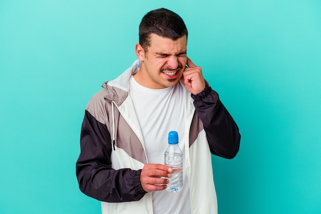 Agua potable del hombre caucásico deportivo joven aislado en azul que cubre los oídos con las manos.