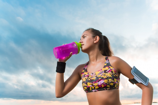 Agua potable de la muchacha apta de los jóvenes en la playa después del entrenamiento de la mañana