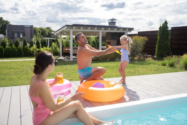 Agua sobre papá. Hija divertida salpicaduras de agua sobre papá mientras se divierte con los padres cerca de la piscina