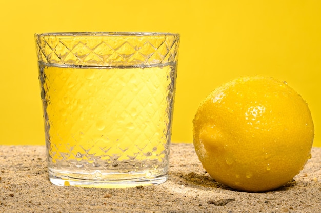 Agua en vaso y limón sobre arena contra fondo amarillo