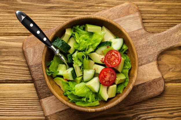 Aguacate casero saludable, pepino, ensalada de hojas verdes en un tazón de madera sobre una tabla de cortar sobre fondo marrón rústico de madera. Comida vegetariana, merienda saludable para la dieta y una alimentación limpia. Vista superior