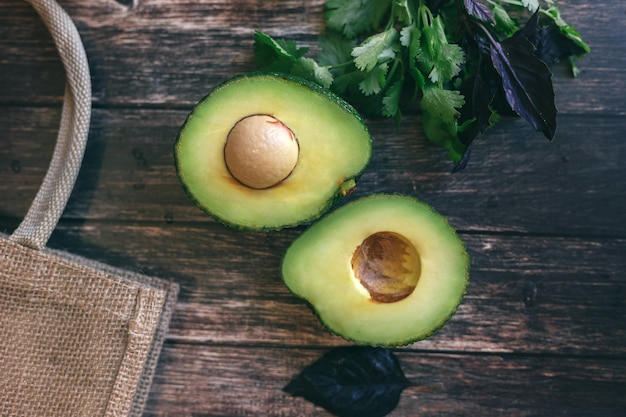 Aguacate cortado por la mitad sobre una mesa de madera, cilantro y albahaca junto a una bolsa de paja. concepto de nutrición adecuada y saludable, vegetarianismo.