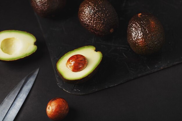 Aguacate Hass orgánico en rodajas y entero con un cuchillo sobre una superficie negra. Fuente de grasas esenciales, vitaminas, oligoelementos, betacaroteno y ácidos grasos omega-3.