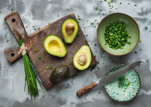 Aguacates frescos cortados en una tabla de cortar de madera con cebollas verdes y un cuenco de hierbas picadas sobre un fondo gris texturizado