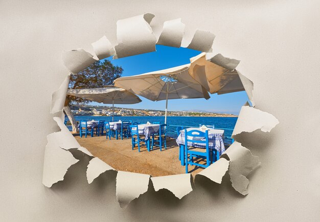 Agujero de papel con imagen de restaurante junto al mar con mesas vacías y sombrillas abiertas listas para turistas.