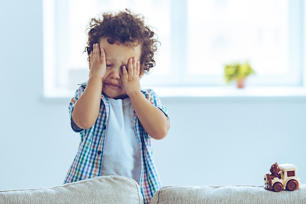 Ah não! Bebezinho africano chorando e esfregando os olhos enquanto está de pé no sofá de casa