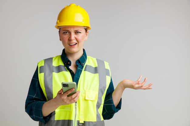 Foto ahnungsloser junger weiblicher bauarbeiter, der sicherheitshelm und sicherheitsweste hält, die handy hält leere hand