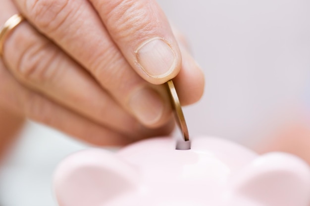 Ahorros, dinero, seguro de anualidad, jubilación y concepto de personas - cerca de la mano de mujer senior poniendo monedas en hucha