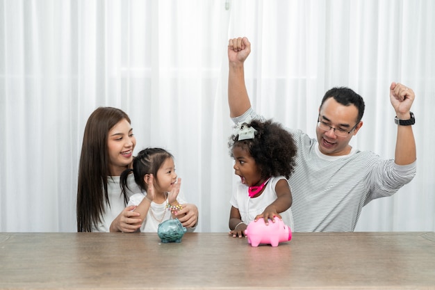 Foto ahorros familiares, planificación presupuestaria, dinero de bolsillo de los niños, familia asiática e hija africana adoptada muestran hucha